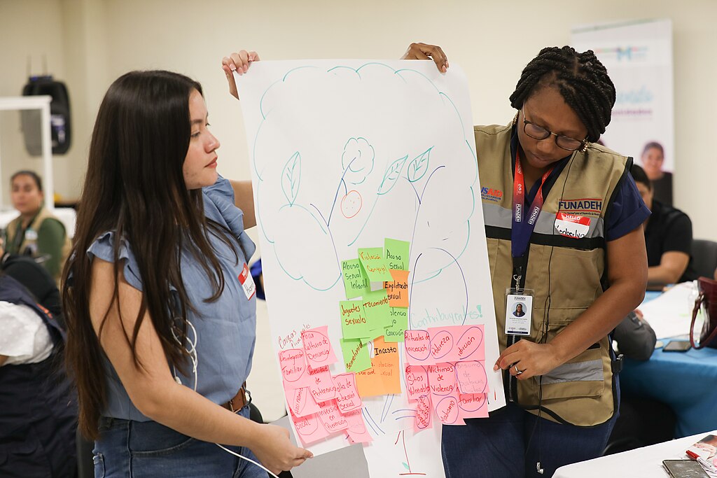 Two women present a large paper with a hand-drawn tree diagram covered in colorful sticky notes during an indoor seminar.