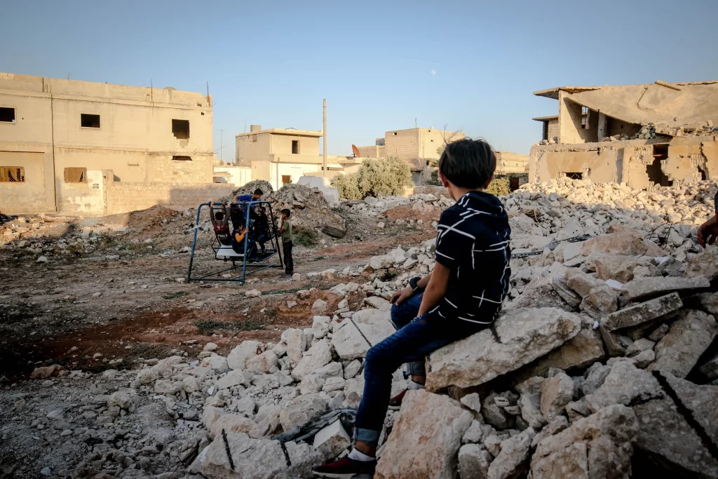 Children play on a swing in a war-torn area of Idlib, showcasing resilience amidst devastation.
